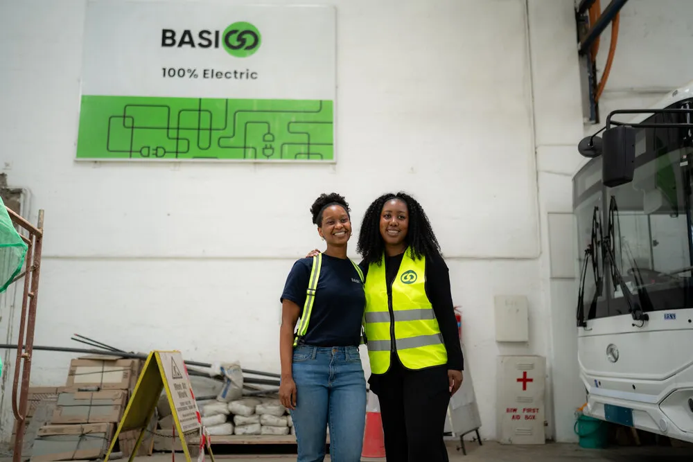 Two women posing in front of the BasiGo 100% Electric sign - one in a BasiGo navy t-shirt, the other in a yellow safety vest