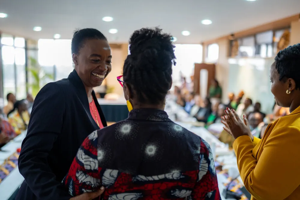 Three women in warm interaction - woman in navy blazer laughing, woman in red/navy African print seen from behind, woman in yellow blazer clapping, audience applauding