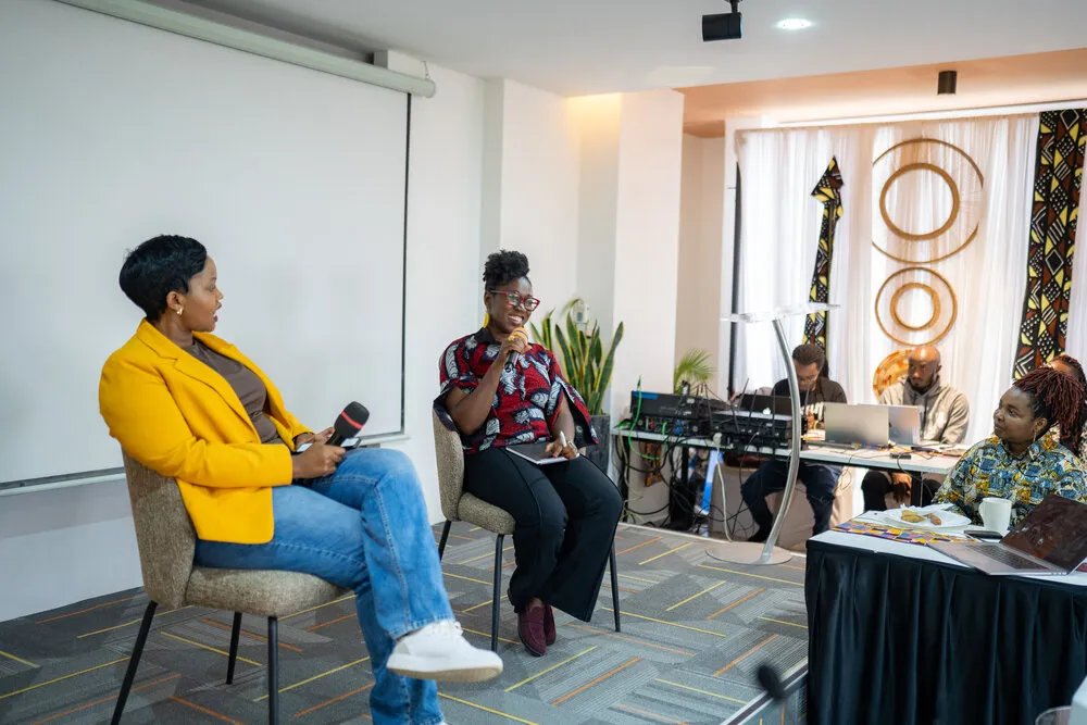 Dr. Joy Buolamwini in red and black African print speaking into a microphone, seated next to a woman in a yellow blazer