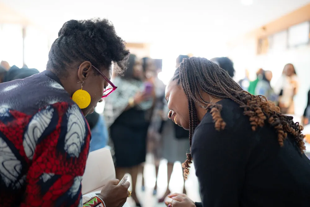 Woman in red/navy African print with red glasses signing something for woman with box braids in black, yellow earrings, networking event in background