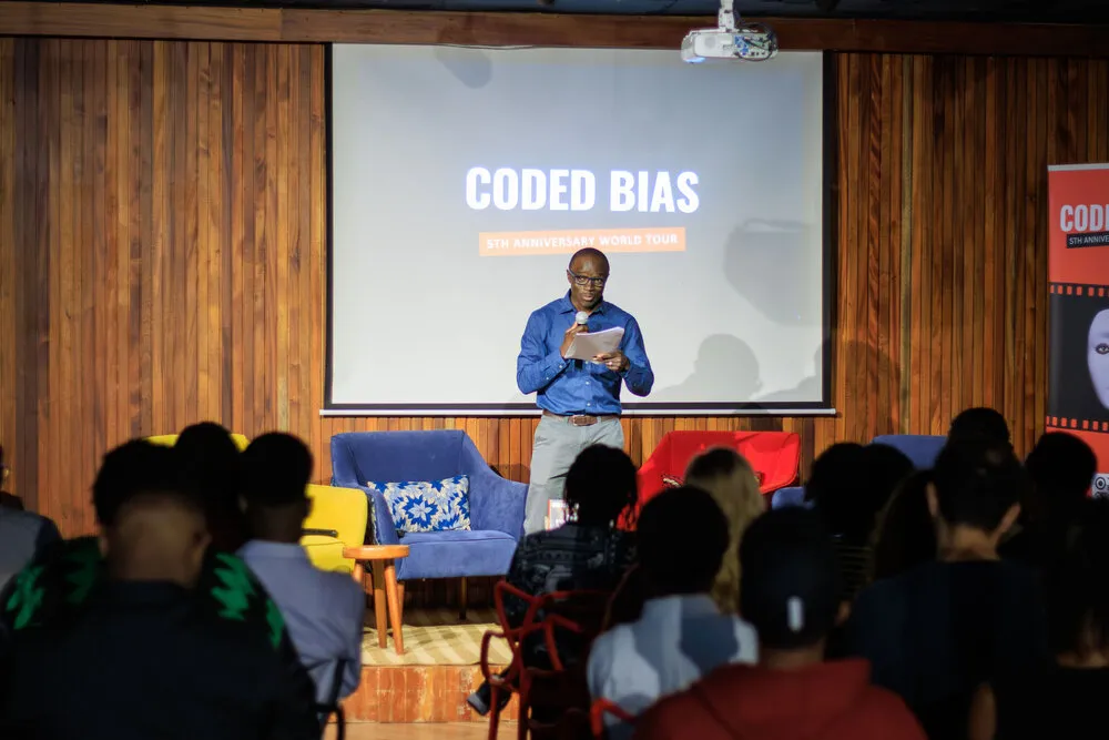 Man in blue addressing seated audience on stage at Baraza, Coded Bias 5th Anniversary on screen, colorful armchairs, wooden panels