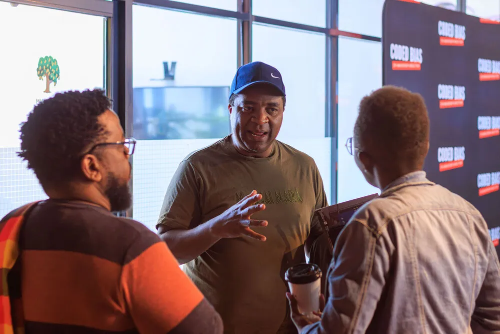 Three men in animated conversation - one in orange/brown stripes with glasses, one in olive Masai Mara tee and Nike cap gesturing, one in denim holding coffee