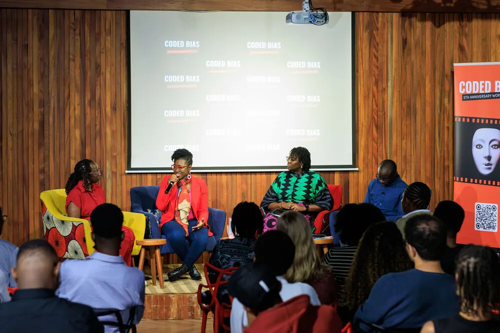 Wide panel shot with audience - four panelists on colorful chairs, woman in red blazer speaking, Coded Bias logos on screen, diverse audience watching