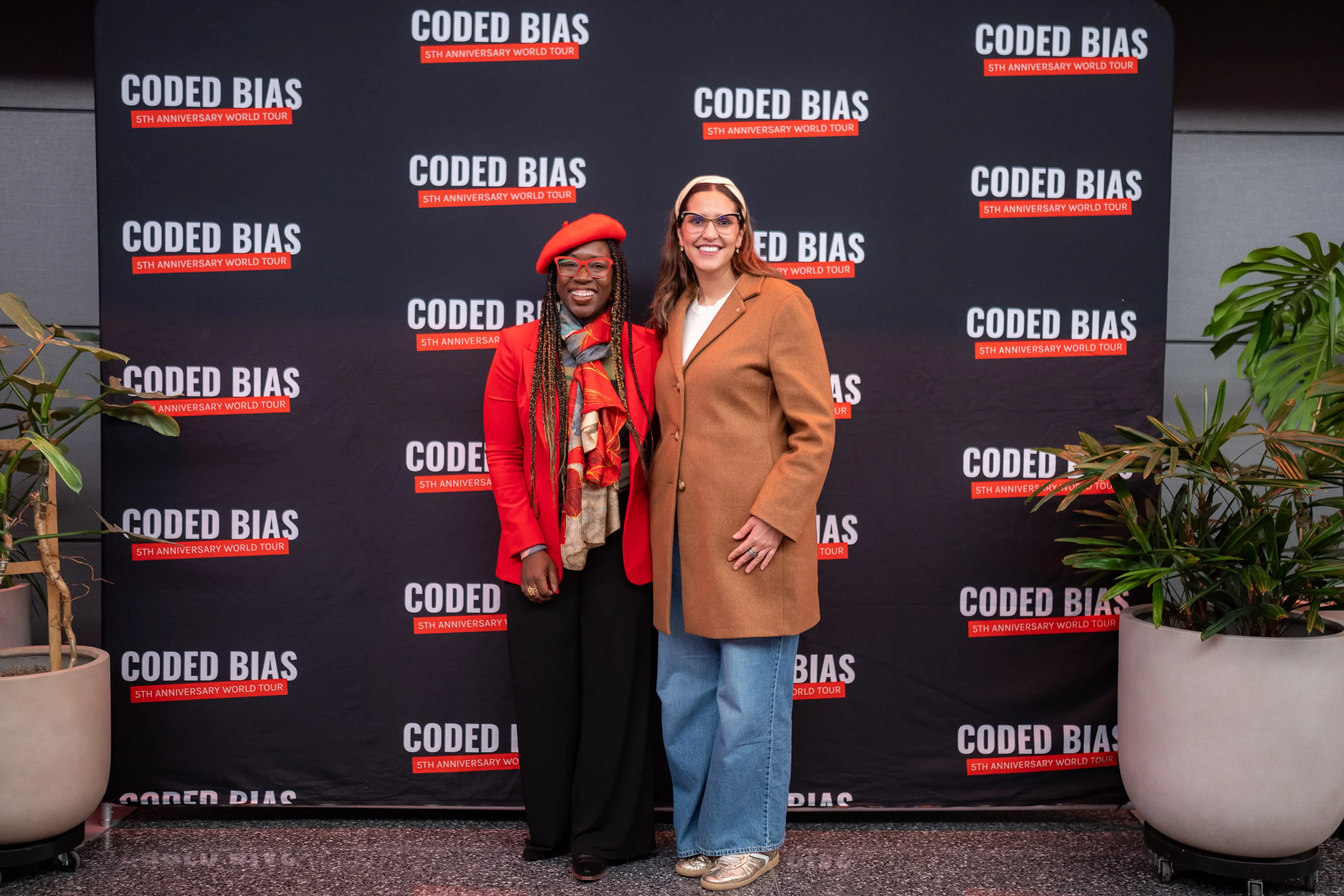 Two women posing together in front of the Coded Bias step-and-repeat banner flanked by potted plants