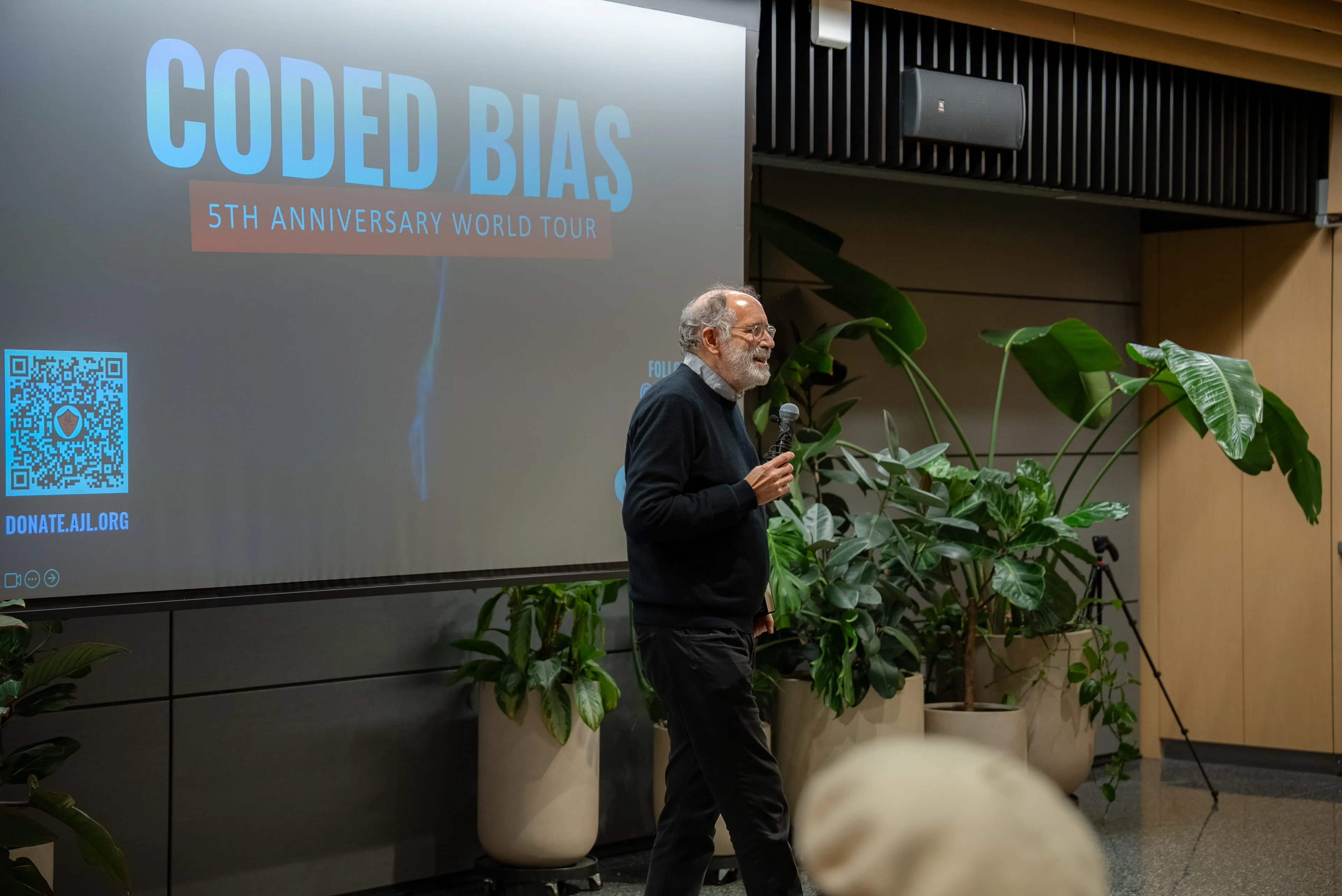 Speaker on stage near the Coded Bias 5th Anniversary World Tour projection screen surrounded by tropical plants