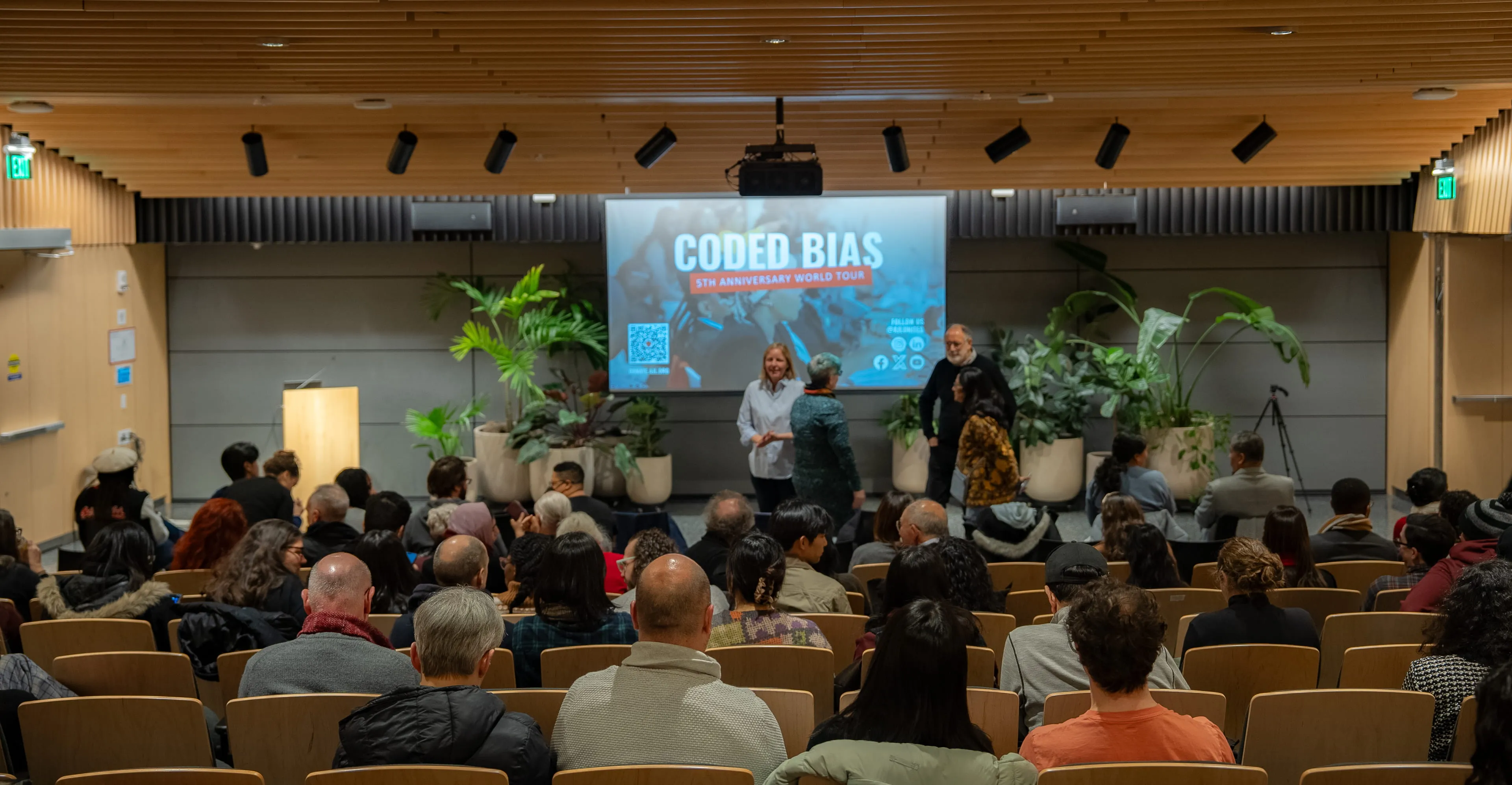 Wide shot of the auditorium with speakers on stage and the Coded Bias 5th Anniversary World Tour slide projected behind