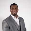 Smiling man wearing a gray suit jacket and white shirt against a plain background.