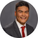 Portrait of a smiling man wearing a dark suit, white shirt, and red tie against a gray background.