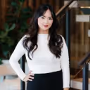 Smiling woman with long black hair wearing a white long-sleeve top with hands on hips in an indoor setting.
