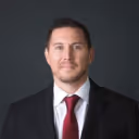 Man with short dark hair wearing a white shirt, red tie, and dark suit jacket against a dark gray background.