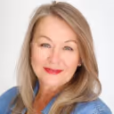 Portrait of a middle-aged woman with blonde hair, smiling and wearing red lipstick against a white background.