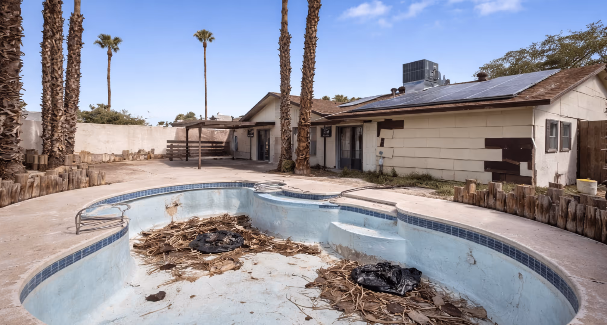 Empty, neglected backyard pool filled with dried leaves and debris, with a single-story house in the background showing patches of repair.