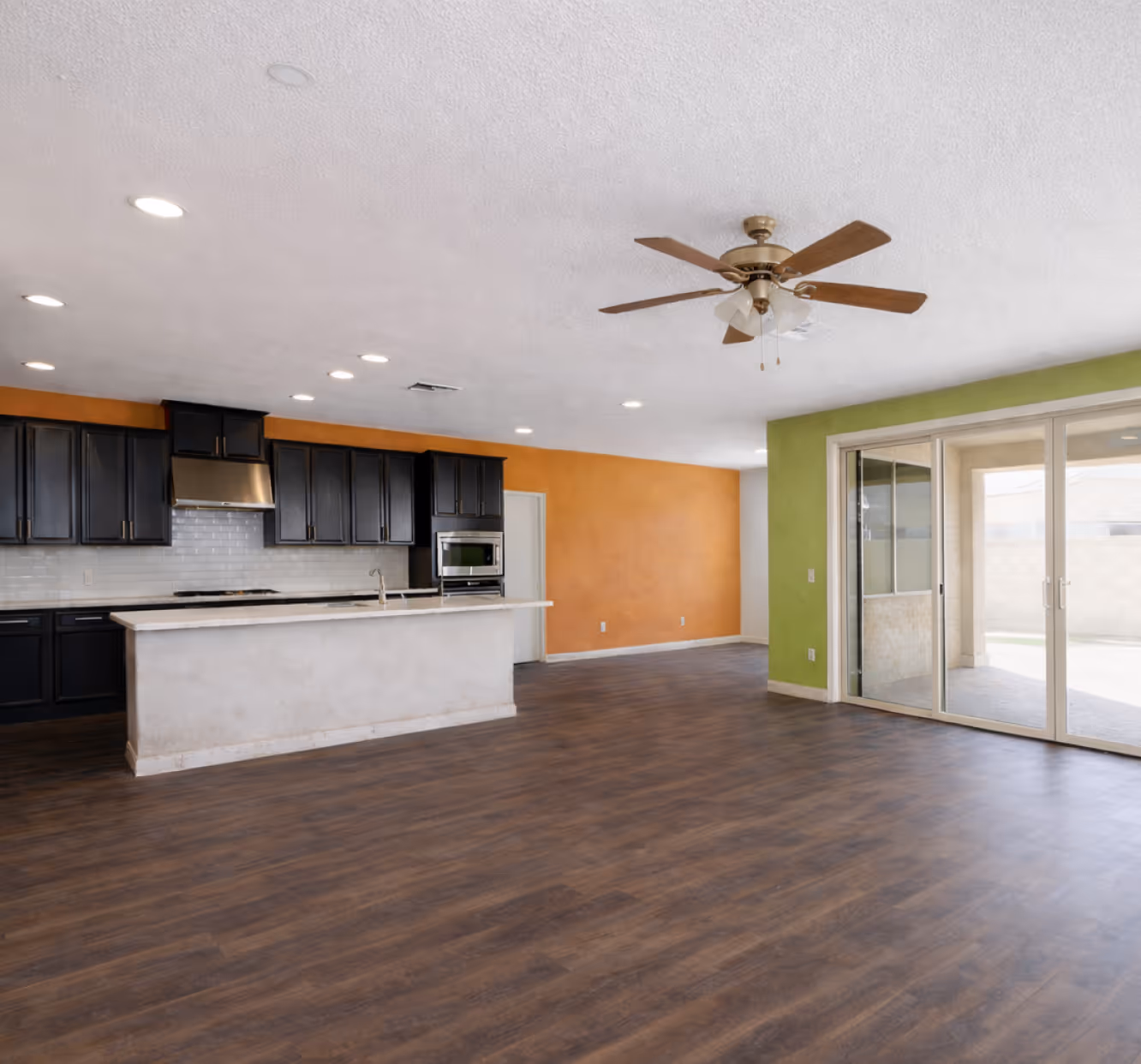 Spacious open-concept kitchen and living area with dark wood cabinets, a white island, orange and green walls, ceiling fan, and sliding glass doors.