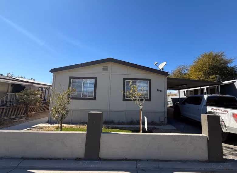 Small beige manufactured home with two windows, small trees in front, a concrete fence, and a white pickup truck parked under a carport.