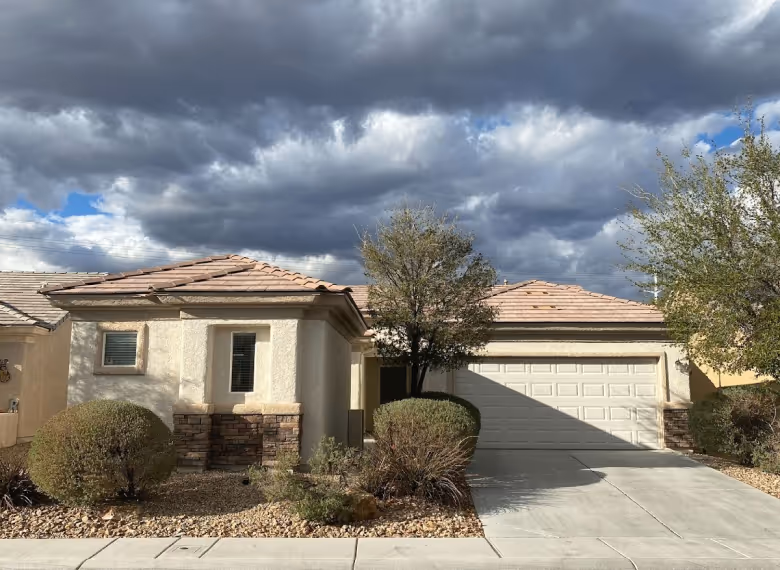 Single-story beige house with tiled roof, two garage doors, trimmed bushes, and ominous dark clouds overhead.