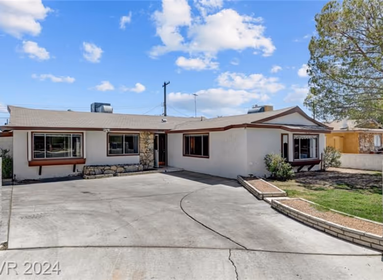 Single-story beige house with a stone accent near the entrance, large concrete driveway, and a tree on the right under a blue sky with clouds.