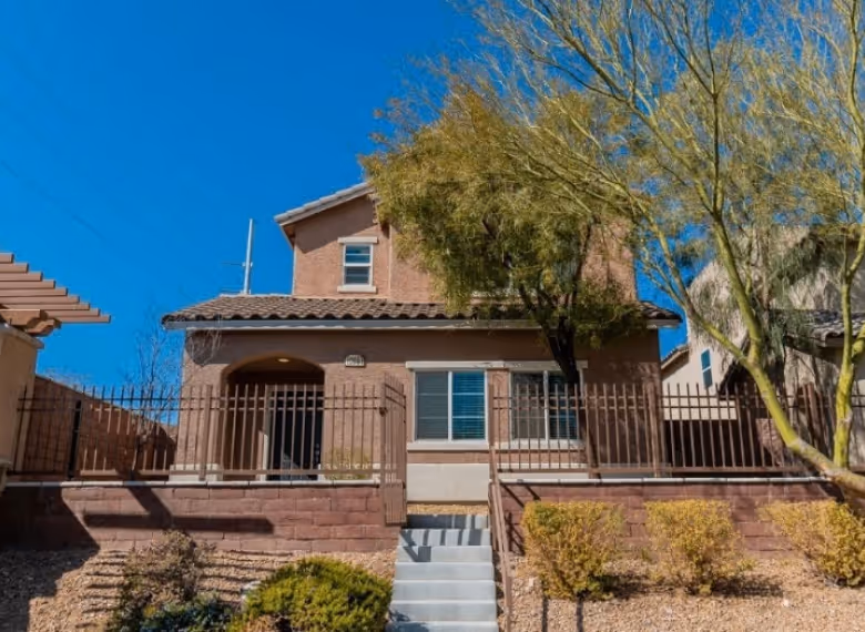 Two-story stucco house with tiled roof, front steps leading to a gated porch, and desert landscaping including bushes and a tree.
