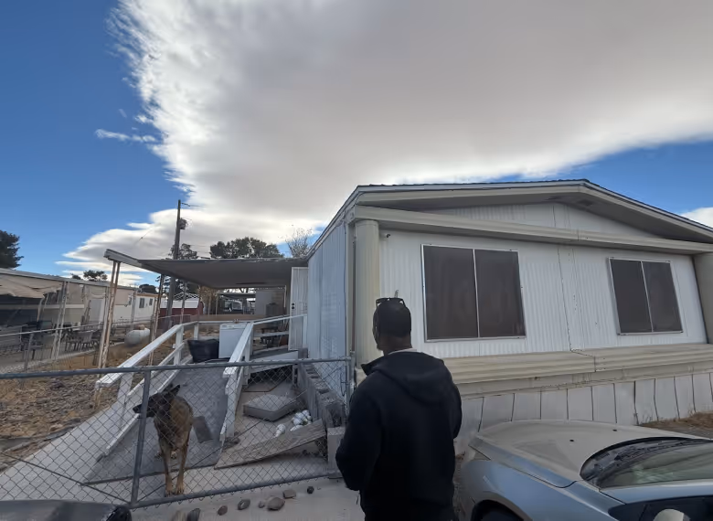 Man in black jacket standing near a fenced ramp beside a white mobile home under a large cloudy sky.