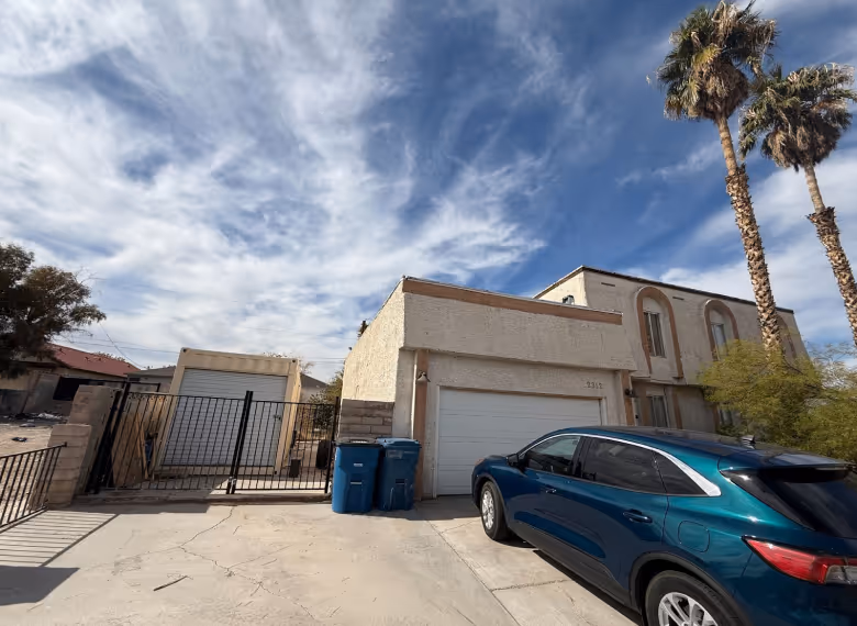 Driveway with a teal SUV parked in front of a beige two-story house with a garage and two palm trees under a partly cloudy sky.