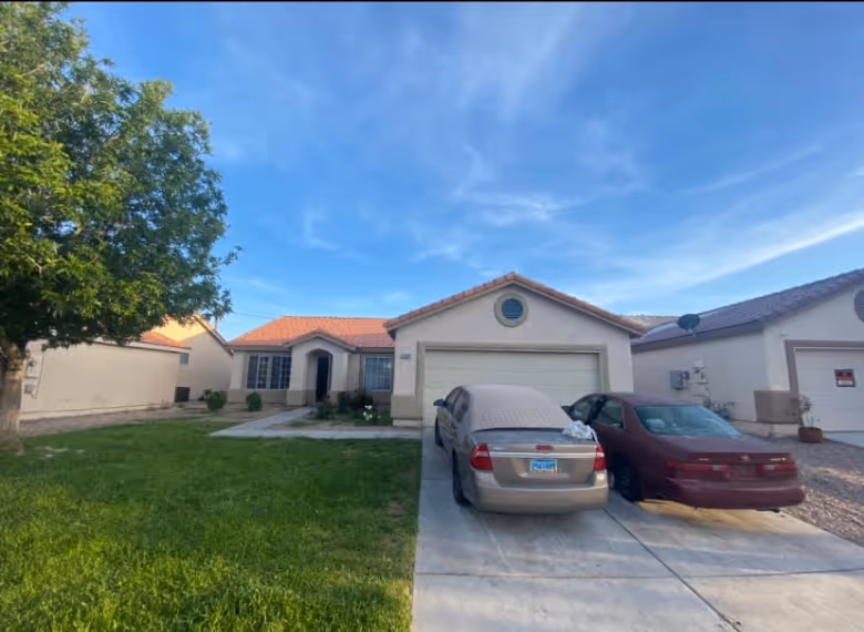 Single-story beige house with a red tile roof, two cars parked in the driveway, a green lawn, and a tree on the left under a clear blue sky.