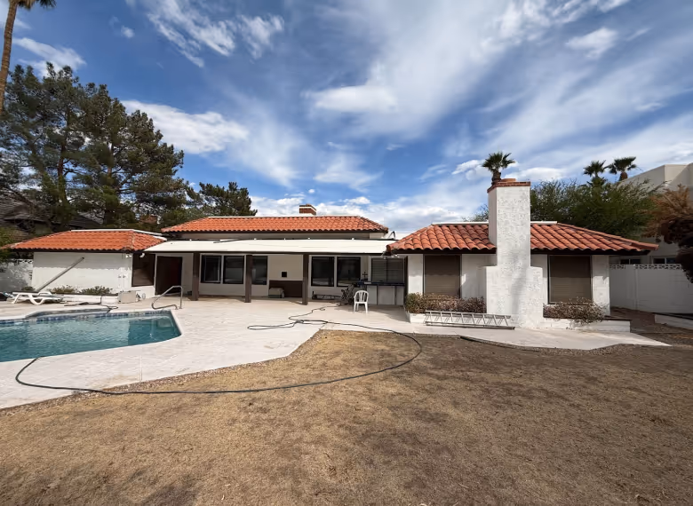 Backyard view of a single-story house with white walls, red tile roof, a swimming pool, and dry grass lawn under a partly cloudy sky.