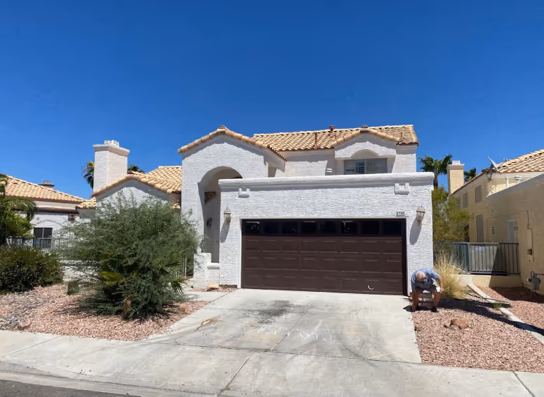 White stucco house with tile roof, brown garage door, and a man crouching by the walkway under clear blue sky.