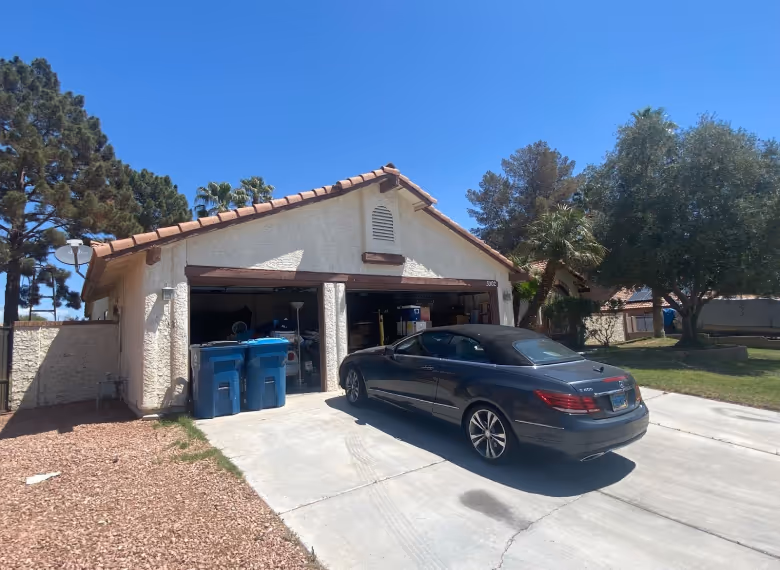 A grey convertible car parked on a driveway in front of a two-car garage with open doors and two blue recycling bins nearby.