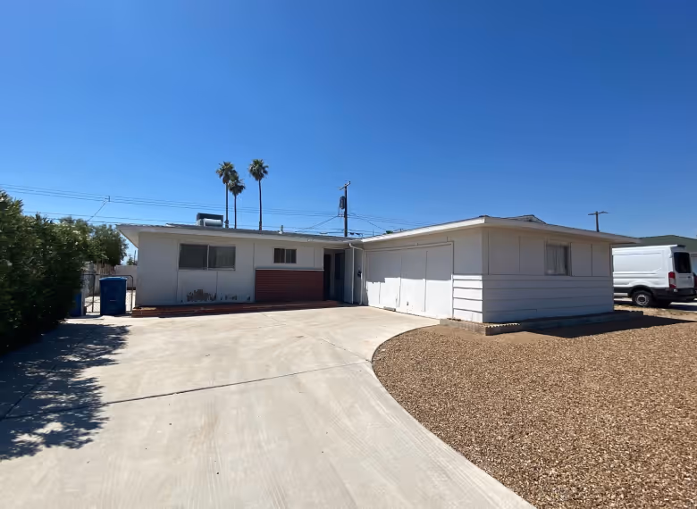 Single-story white house with a large concrete driveway, gravel yard, and three palm trees in the background under a clear blue sky.