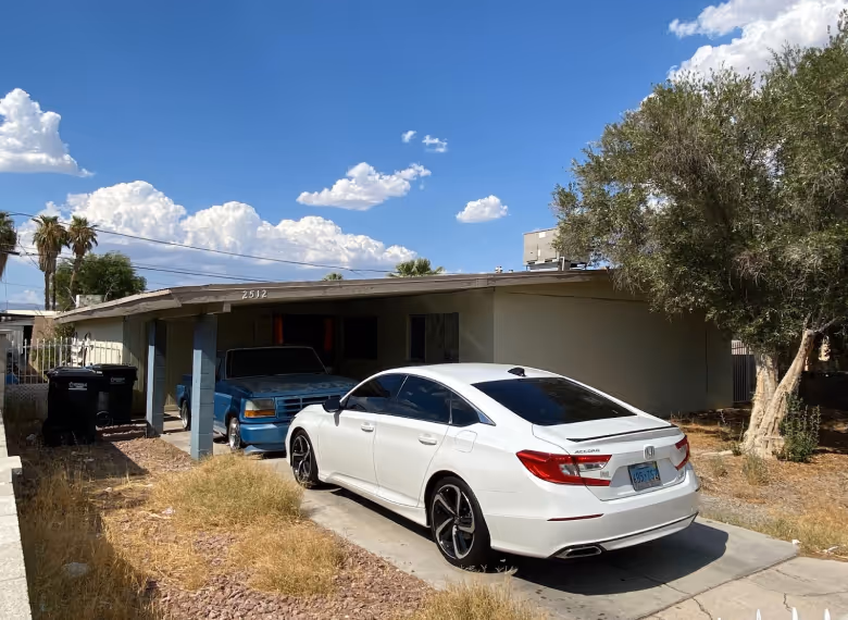 White Honda Accord parked in driveway in front of a single-story house with an attached carport and a blue truck inside it under a partly cloudy sky.