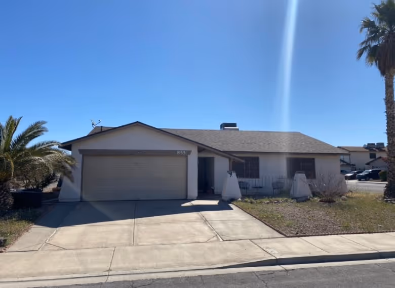 Single-story house with a two-car garage, concrete driveway, and desert landscaping including a palm tree, under a clear blue sky.