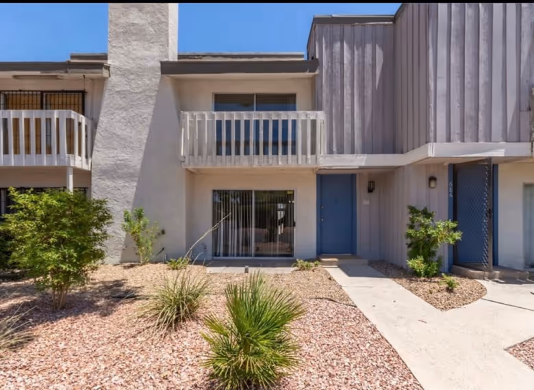 Two-story townhouse with a small balcony, blue door, sliding glass door, and desert landscaping in front.