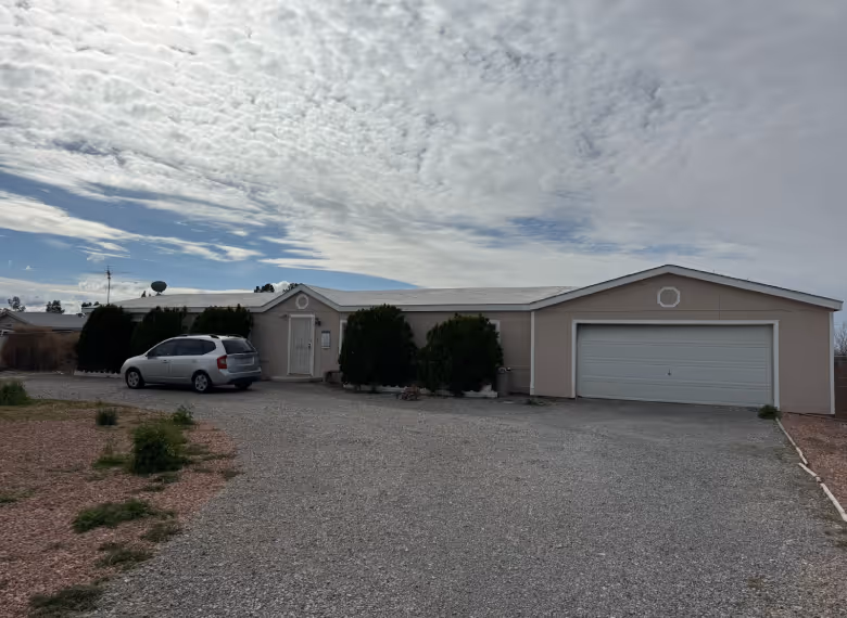Single-story beige house with large gravel driveway, a silver minivan parked near the entrance, and bushes along the front under a cloudy sky.