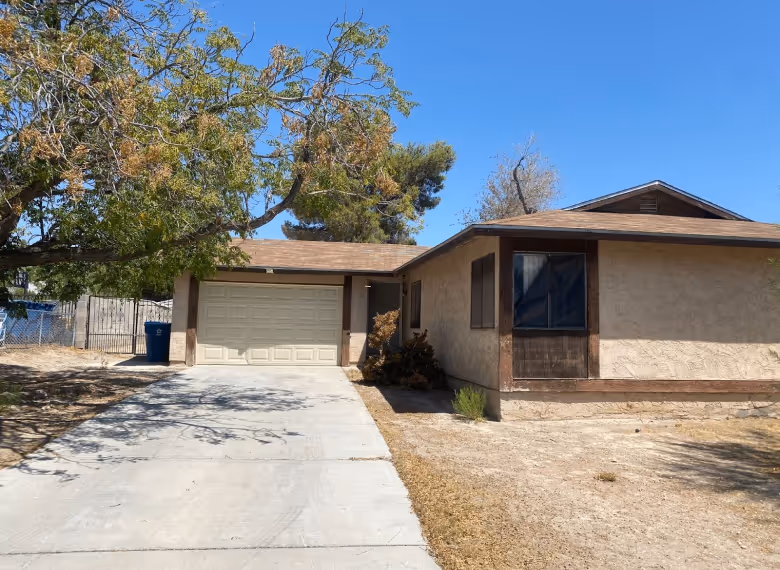 Single-story beige house with brown trim, a closed garage, concrete driveway, and a large tree casting shadows in the front yard.