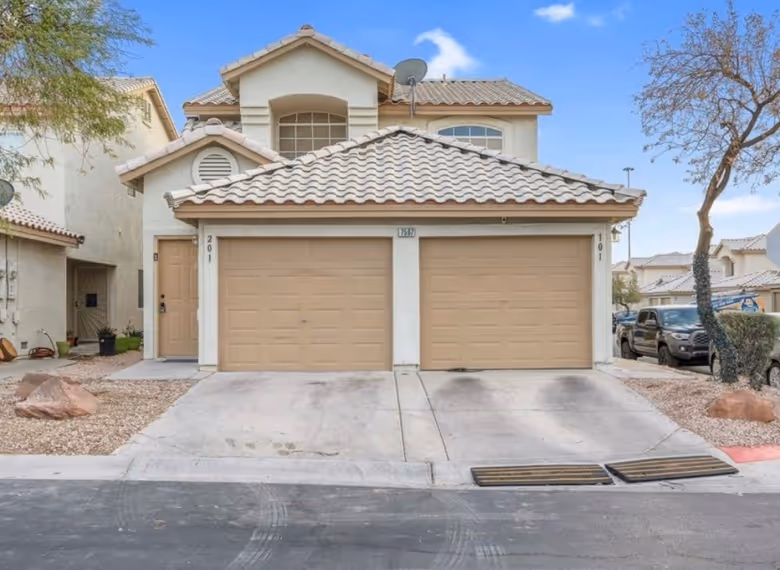 Two-story beige house with tiled roof and double garage doors, a tree on the right, and a driveway in front.