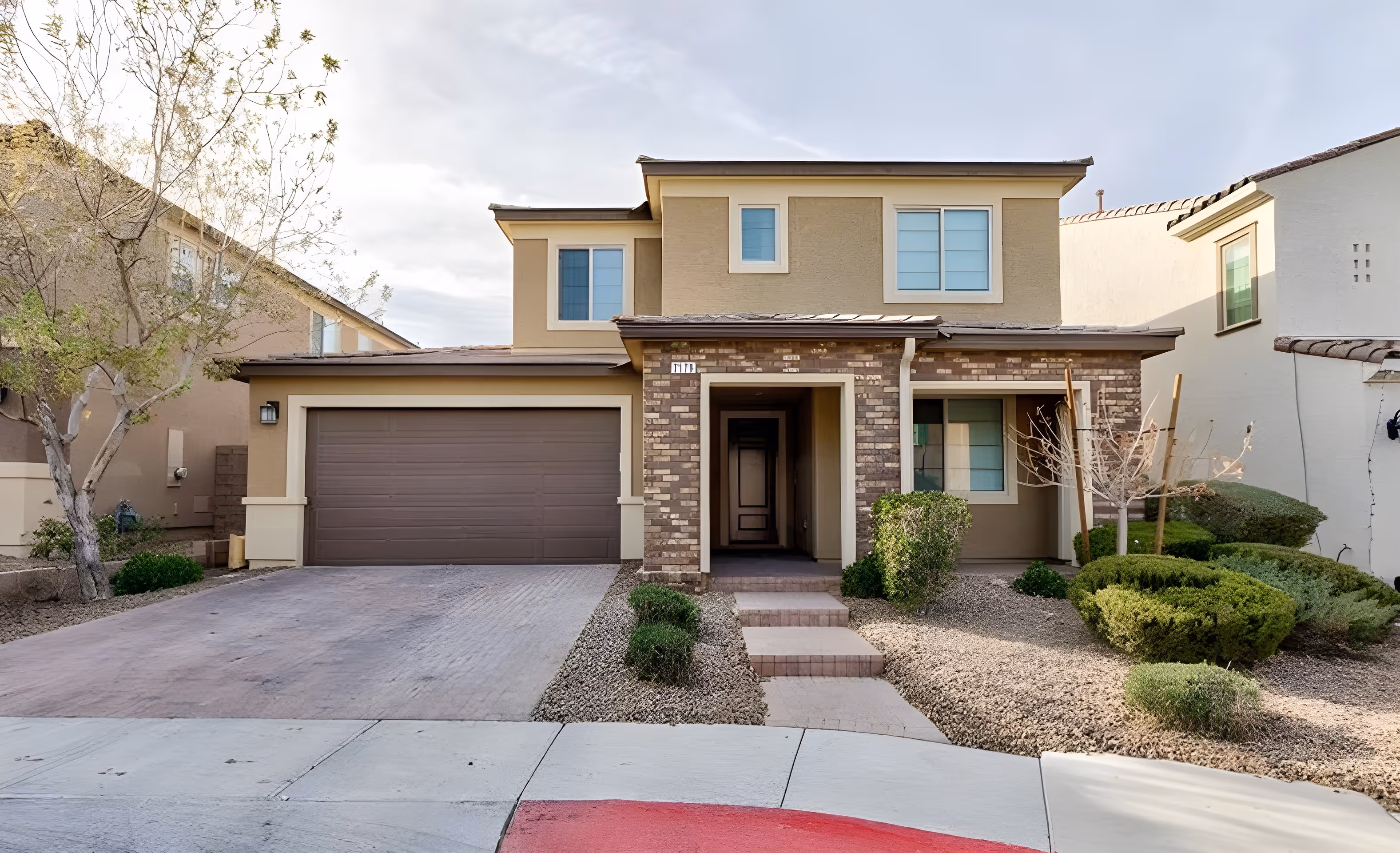 Modern two-story suburban house with a brown garage door, brick entrance, and desert landscaping.