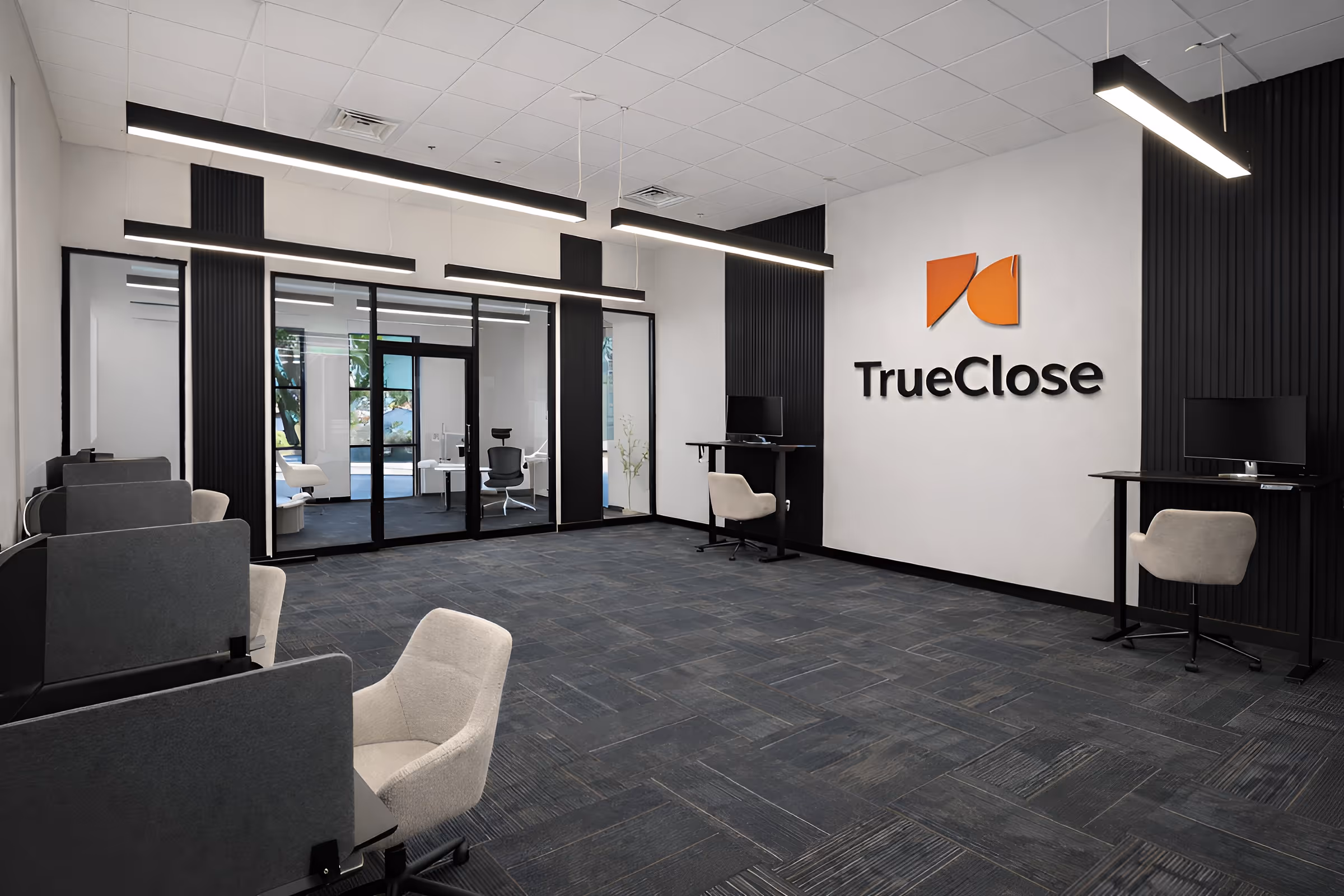 Modern office space with gray carpet, black divider desks, beige chairs, glass wall, and TrueClose logo on white wall.