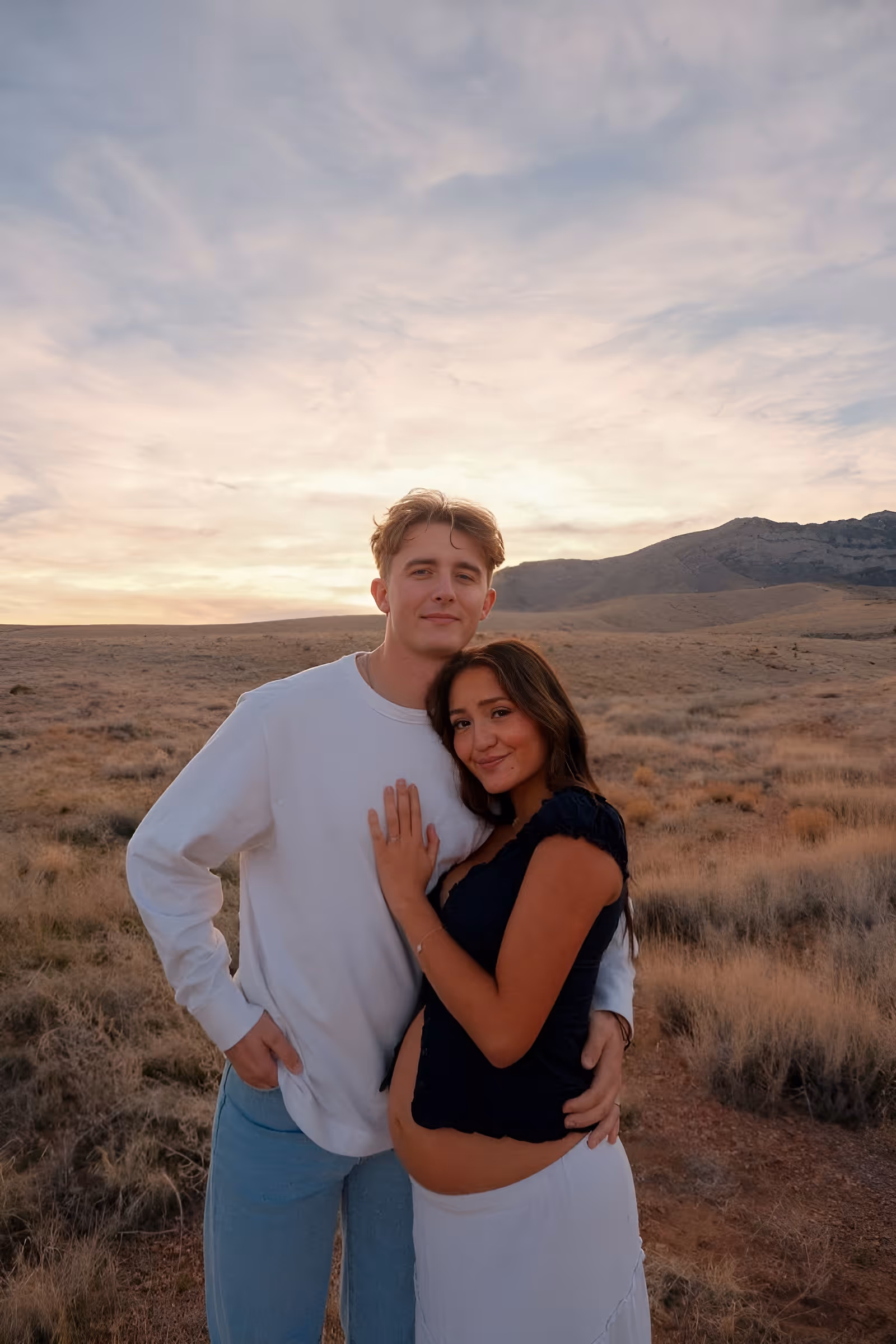Young couple embracing in a dry grassland at sunset, with the woman showing a baby bump.