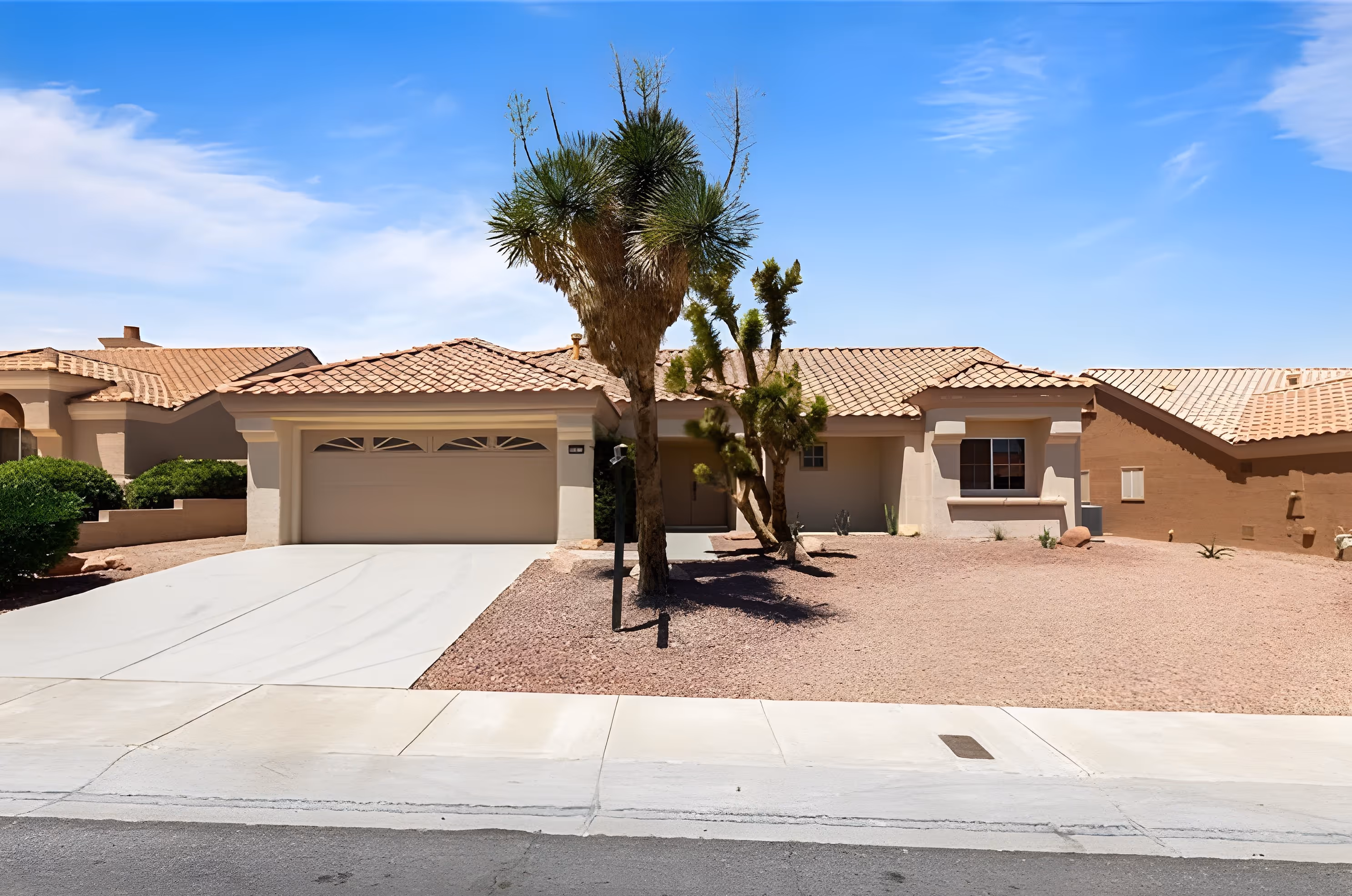 Single-story beige house with a tiled roof, two large trees in the front yard covered with gravel, and a double garage door.
