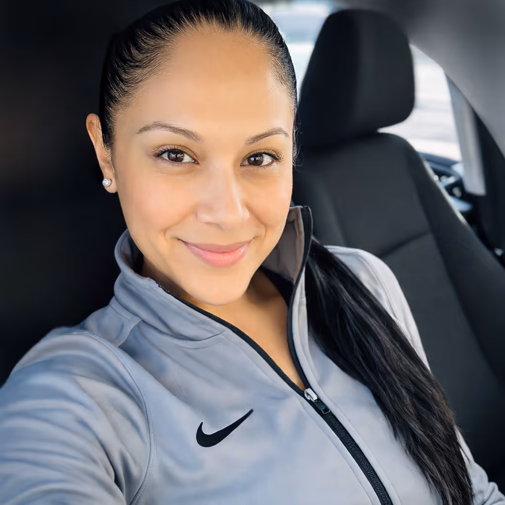 Woman with long dark hair in a gray Nike zip-up jacket smiling inside a car.