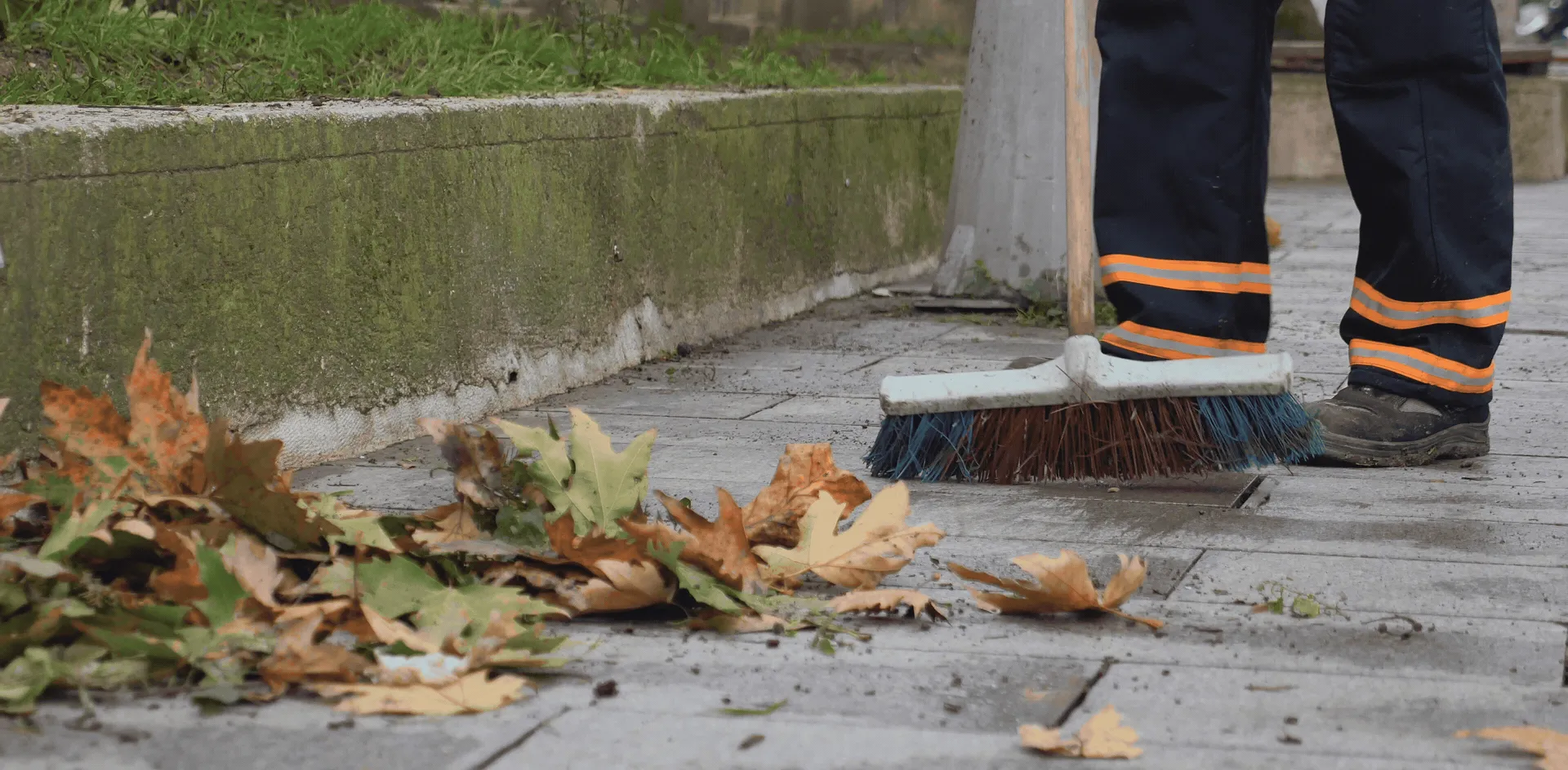 Worker wearing black pants with orange reflective stripes sweeping leaves on a sidewalk.