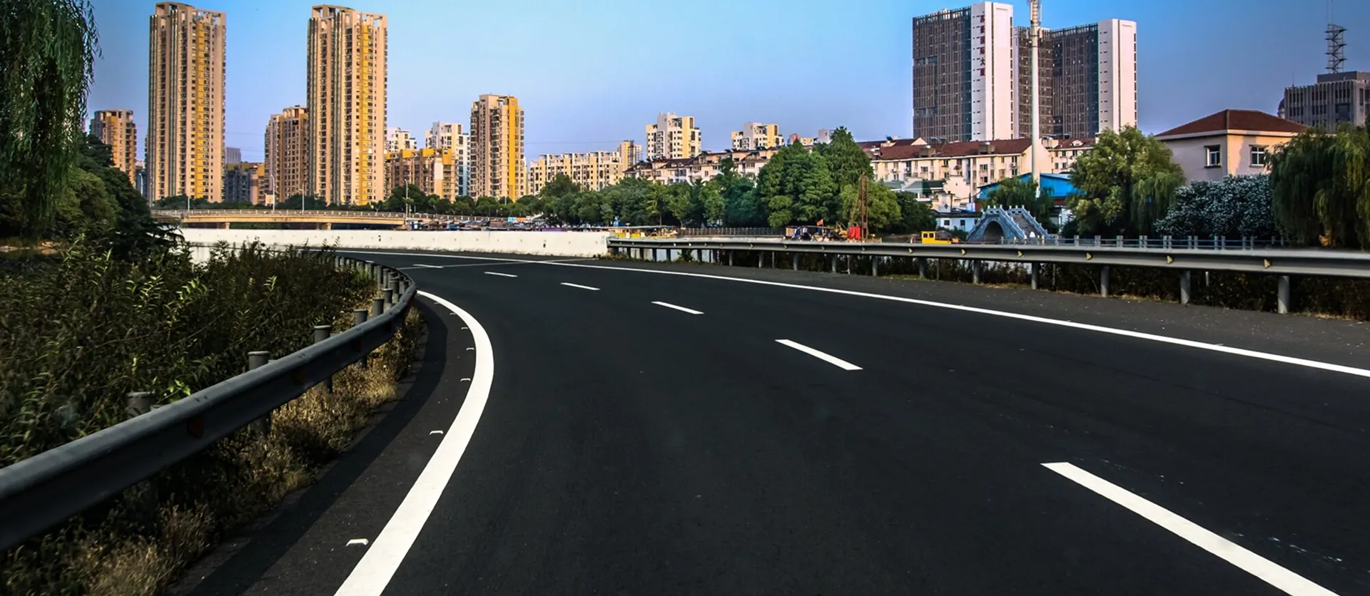 Curved empty highway with guardrails passing through a cityscape with tall residential buildings and trees under a clear blue sky.