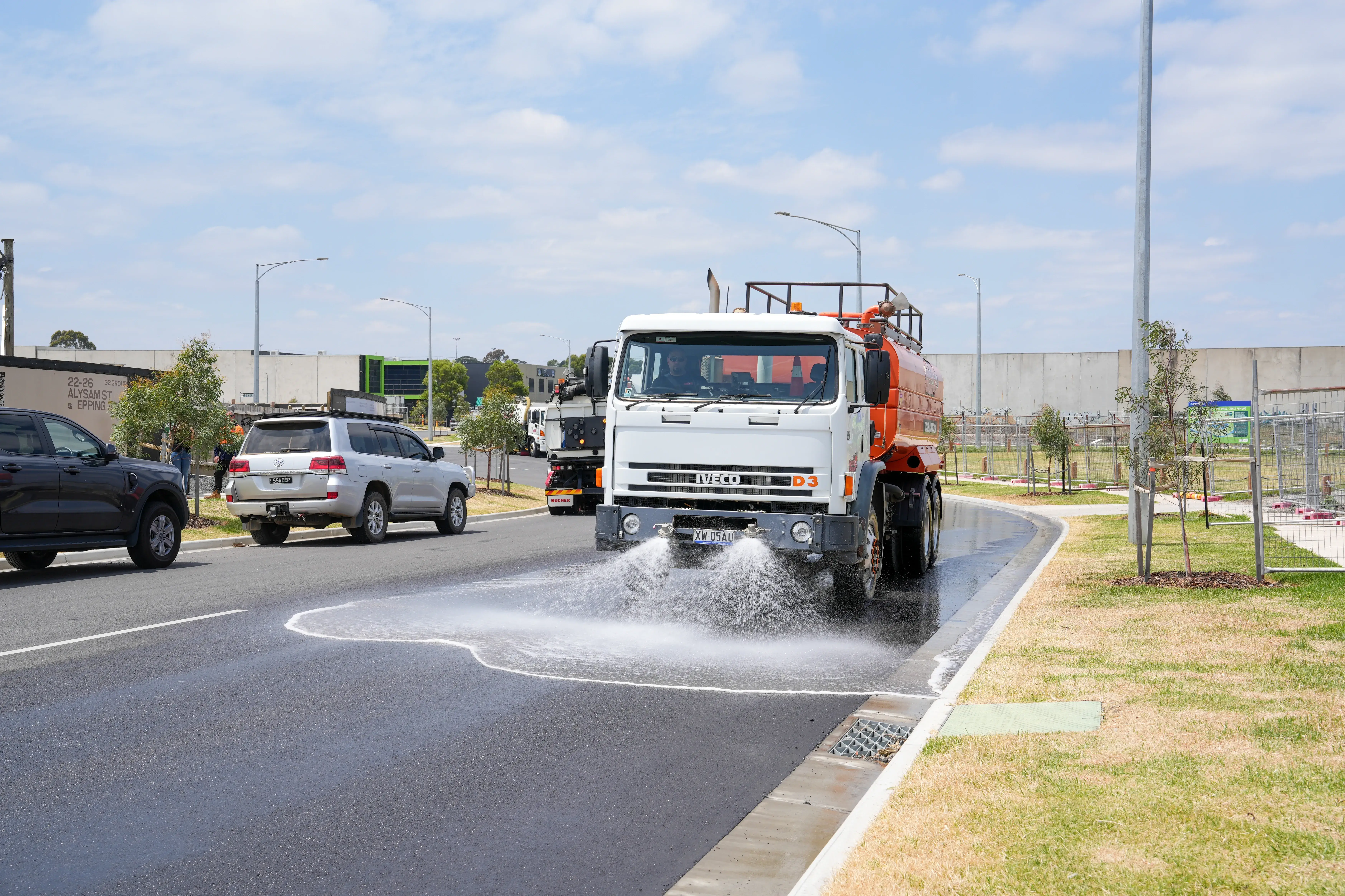 Orange small utility truck driving on a road lined with trees and houses.