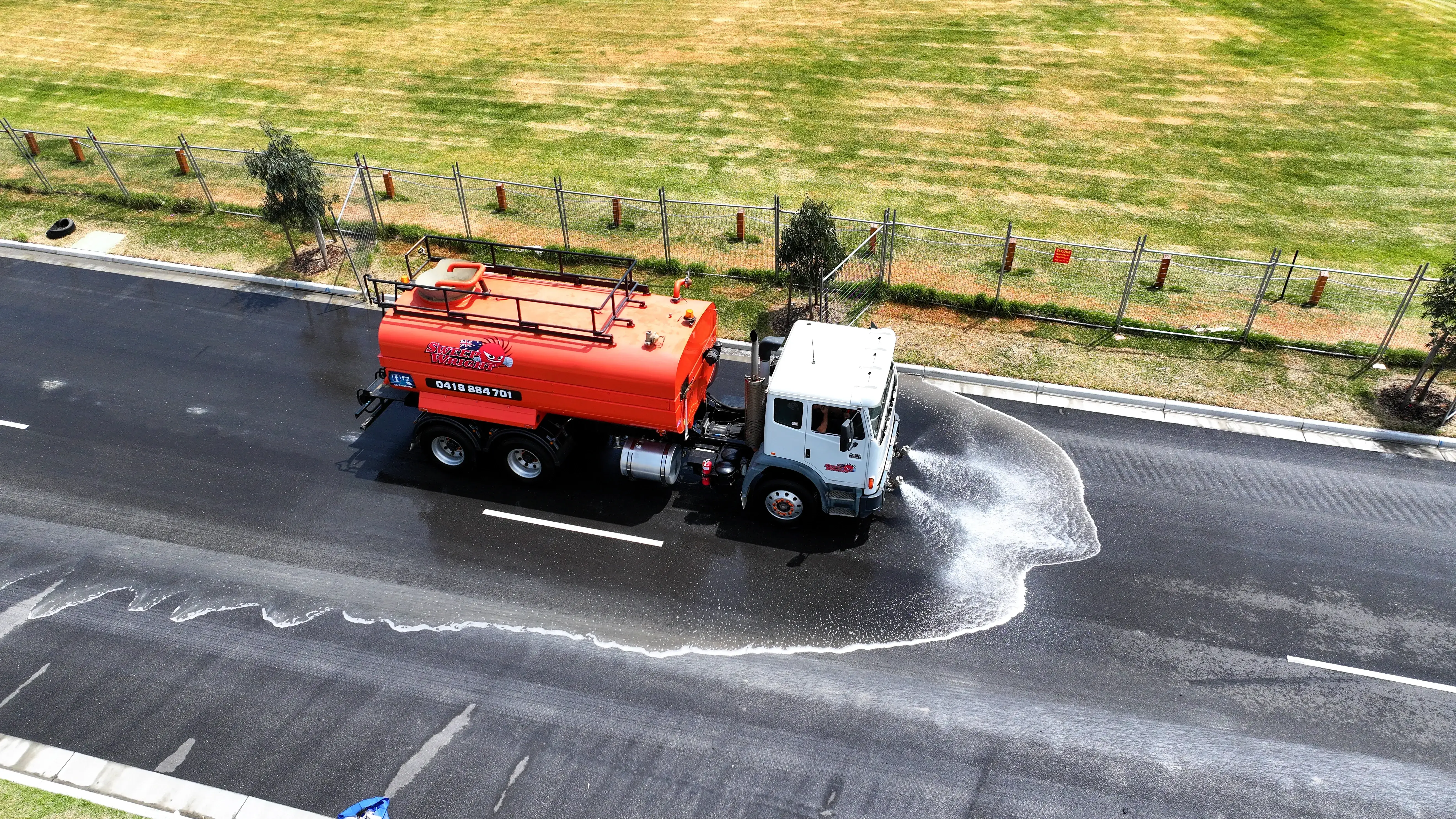 Red and white water truck spraying water on a black asphalt road beside a grass field and chain-link fence.