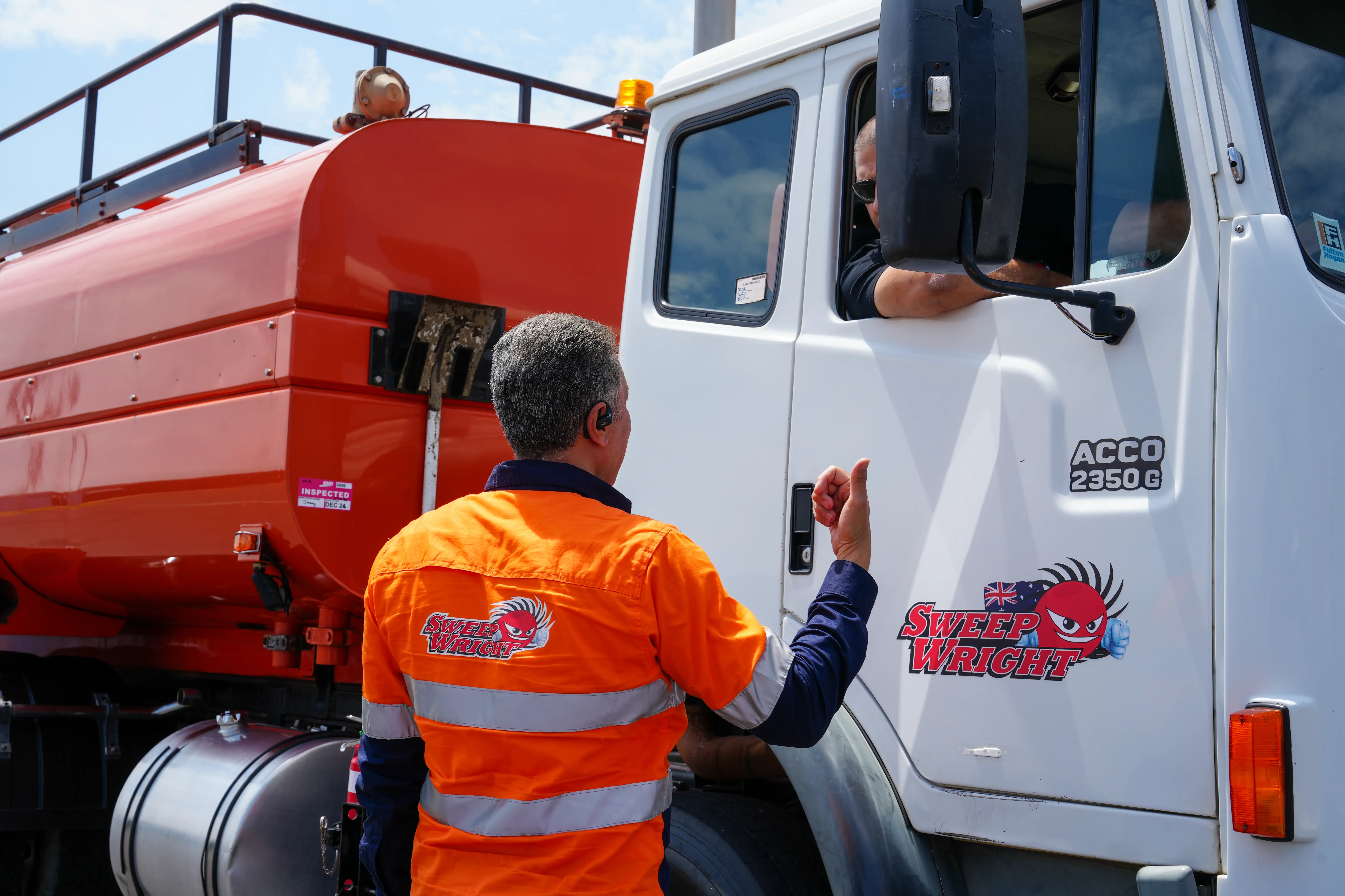 Man in orange safety uniform giving thumbs up to driver in white Sweep WRIGHT truck with orange tank.
