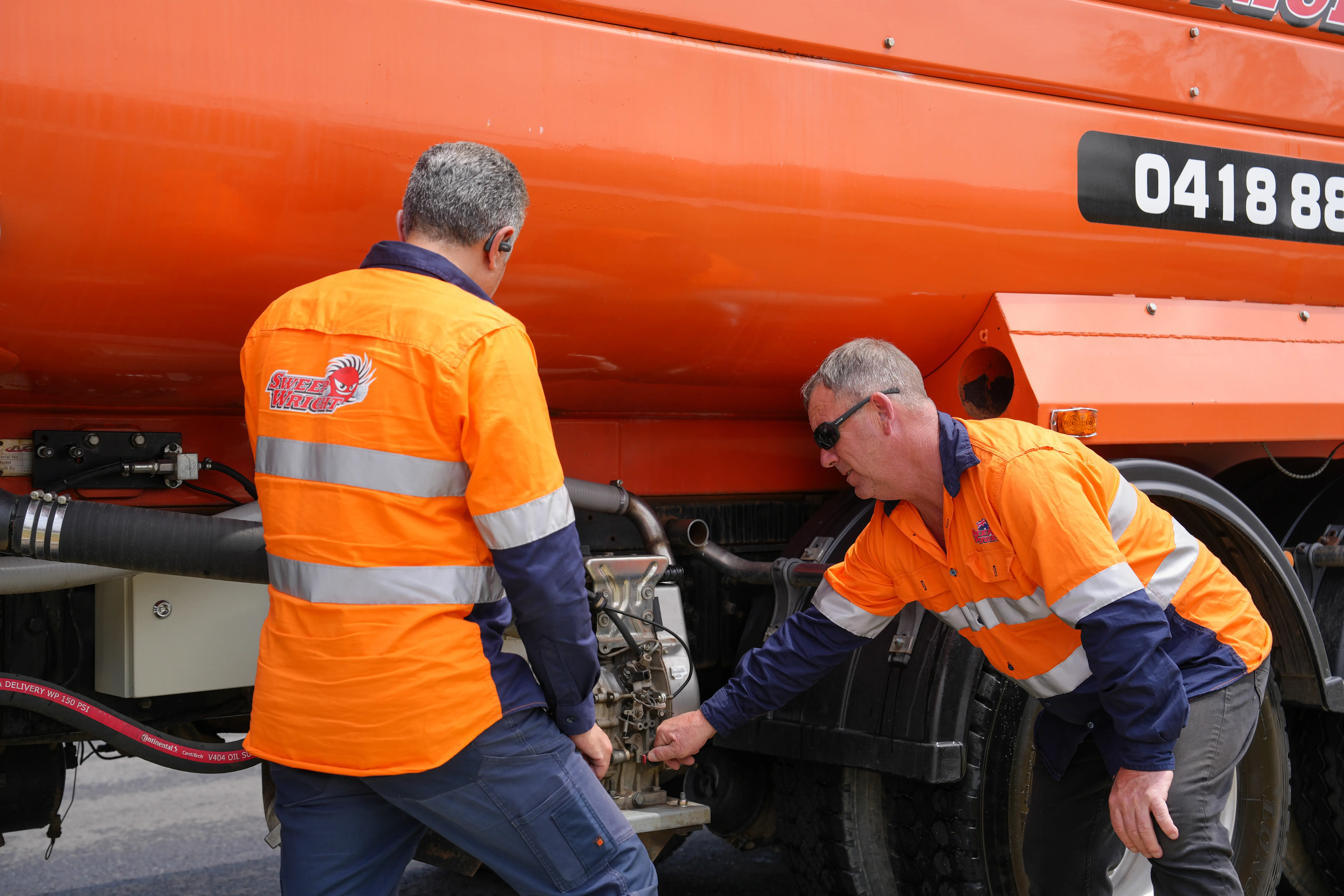Two workers in orange high-visibility jackets inspecting or repairing machinery on an orange industrial vehicle.