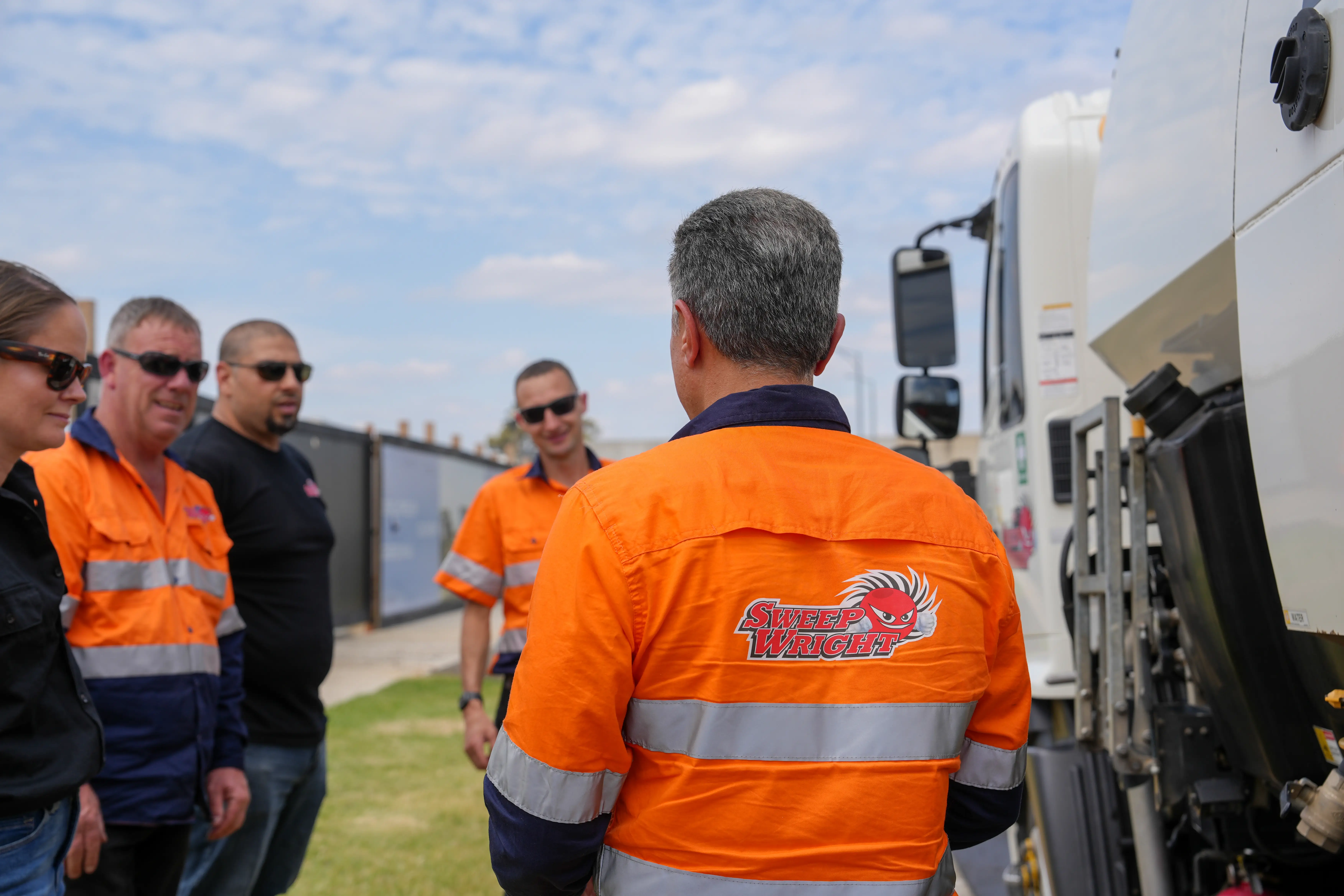 Group of five workers wearing high-visibility orange and black clothing standing outdoors near a white truck, with one worker facing away showing a 'Sweep Wright' logo on the back of his jacket.