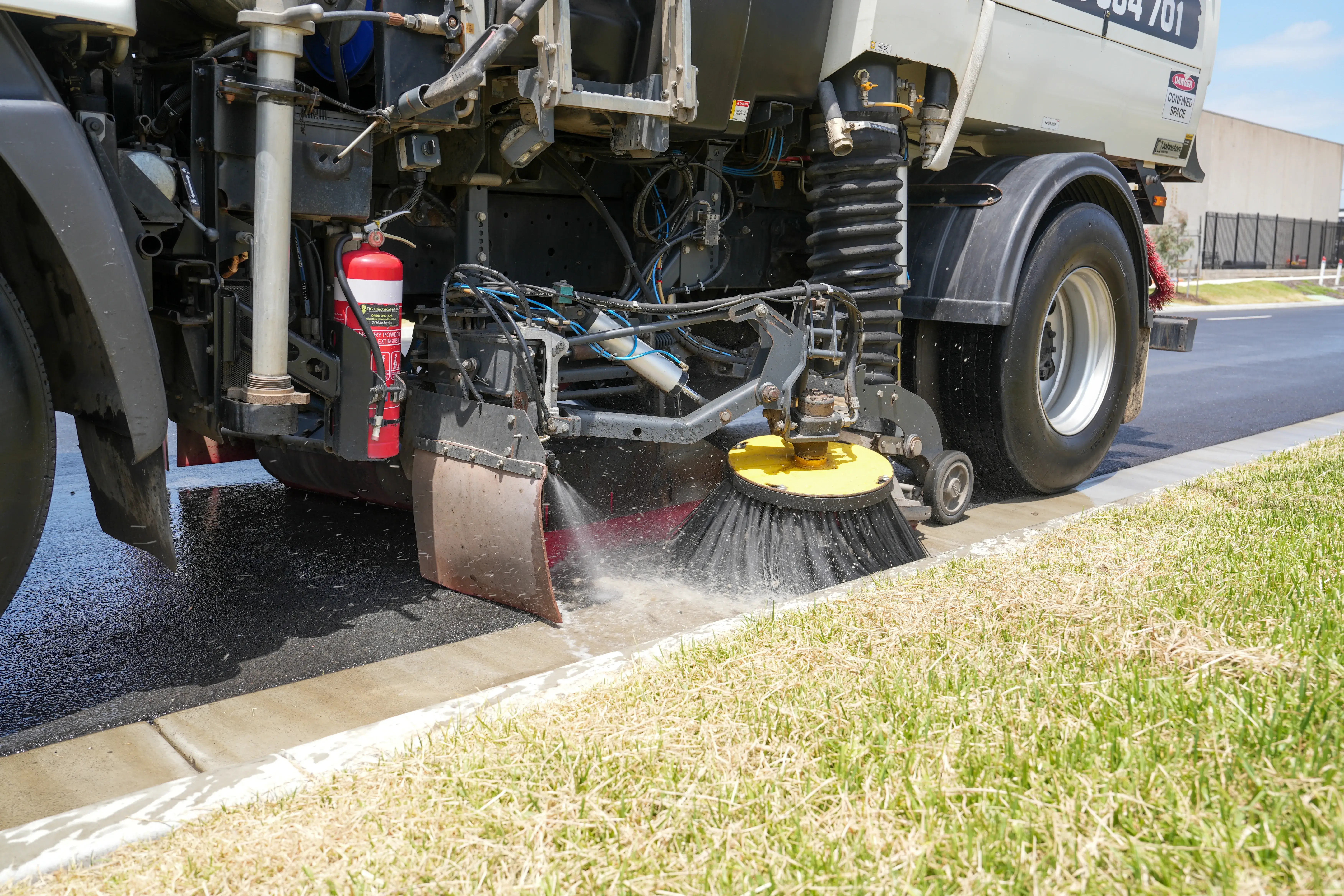Close-up of a street sweeper vehicle cleaning a curb with a rotating brush spraying water.