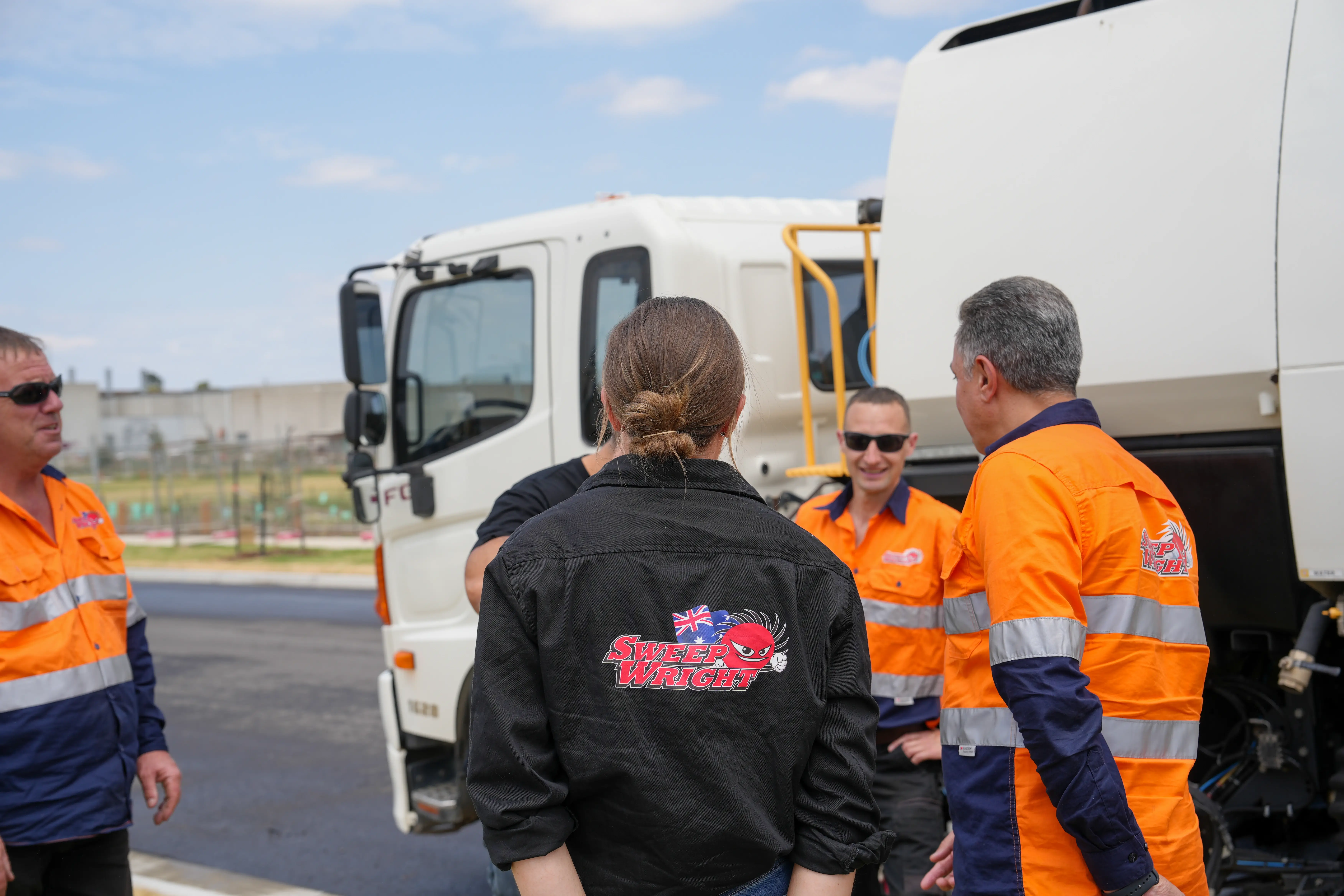 Group of workers in high-visibility uniforms standing and talking near a white street sweeping truck.