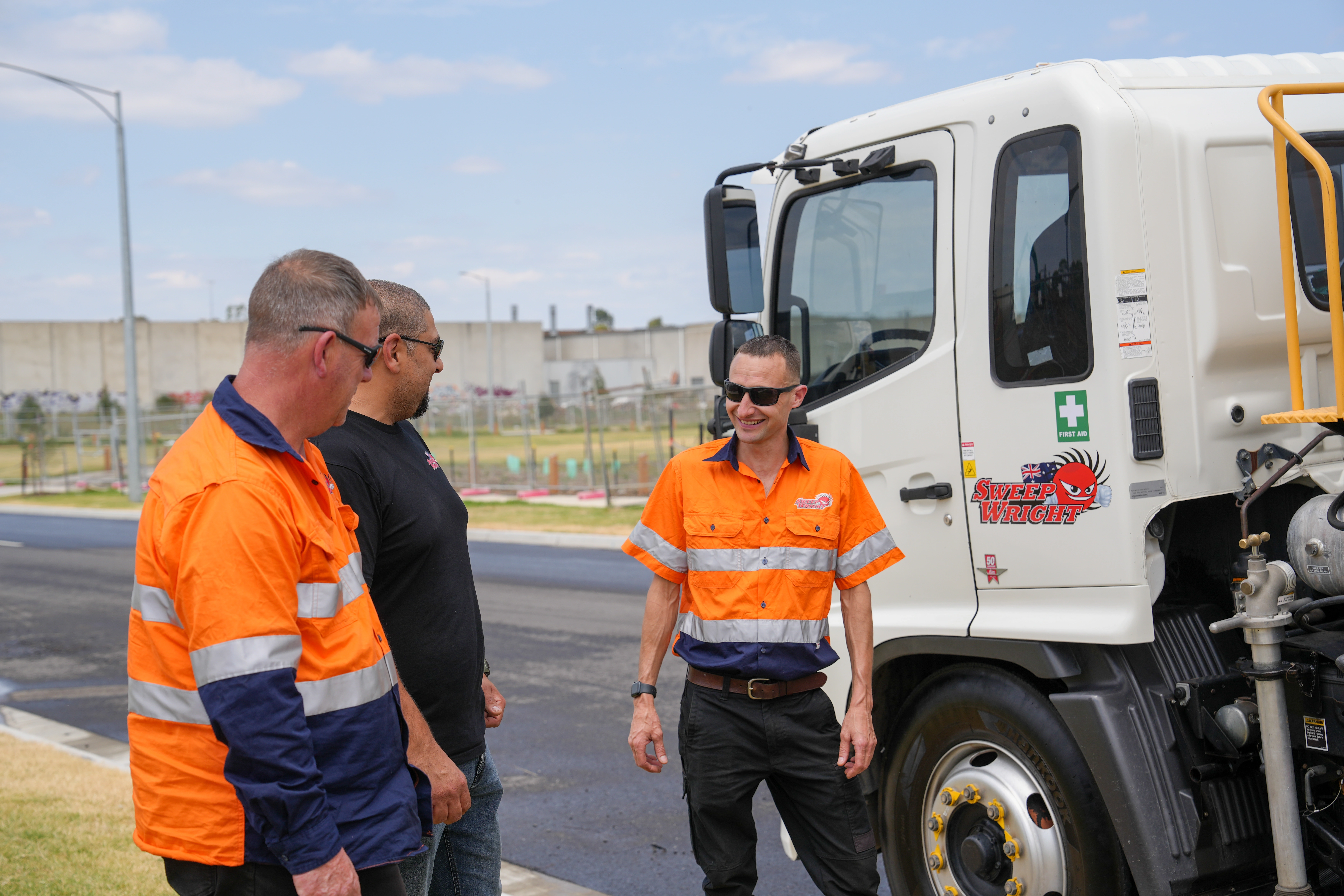 Three men, two in orange high-visibility work shirts and one in a black shirt, standing and talking beside a white Sweep Wright truck.