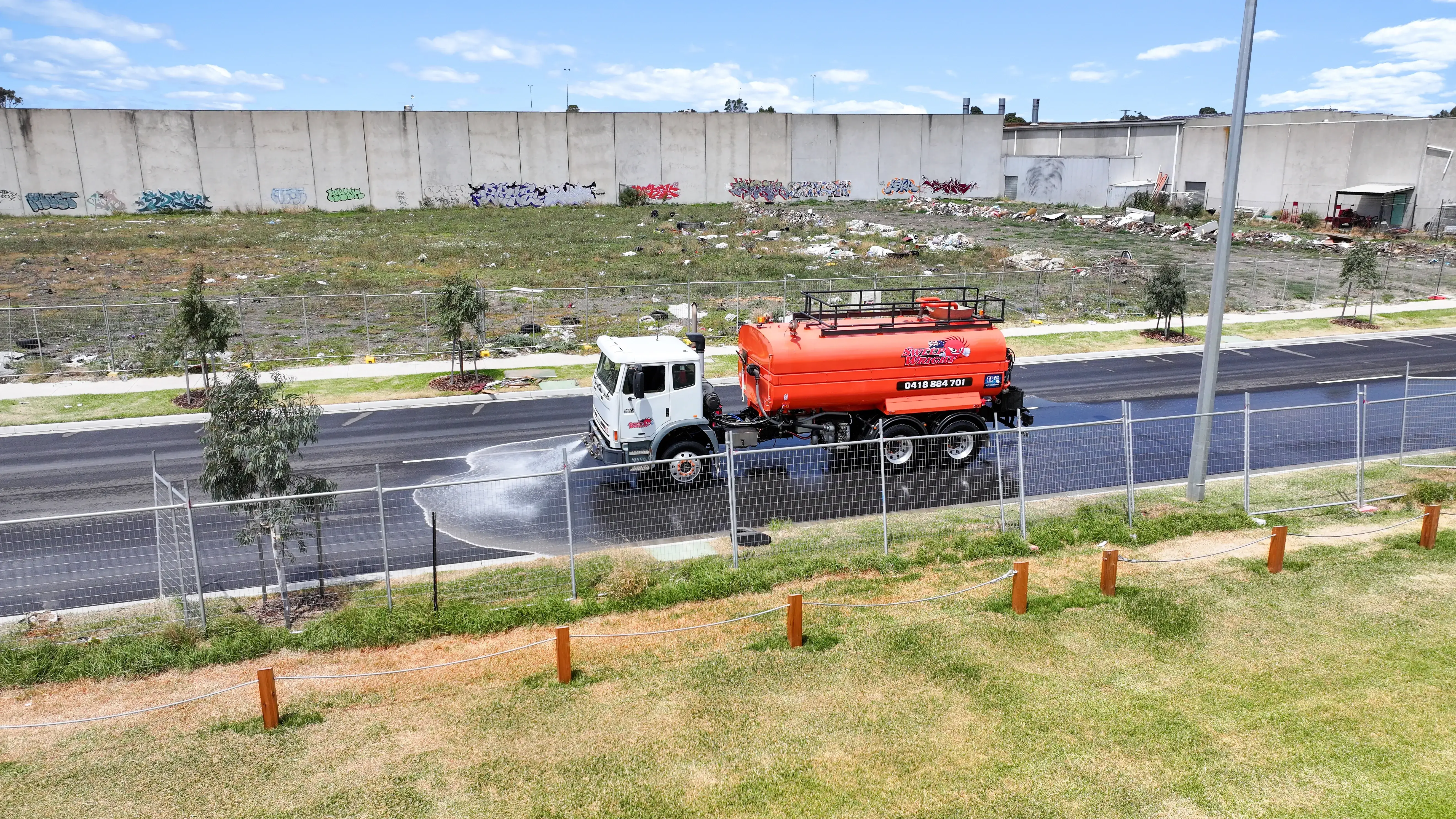 Orange water truck spraying water on a newly paved road beside a fenced grass area and graffiti-covered concrete wall under a blue sky.